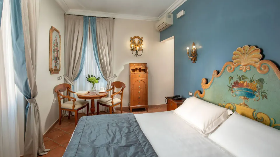 A bedroom with a hand-painted floral headboard, blue walls, a small table with two chairs and pale curtains.