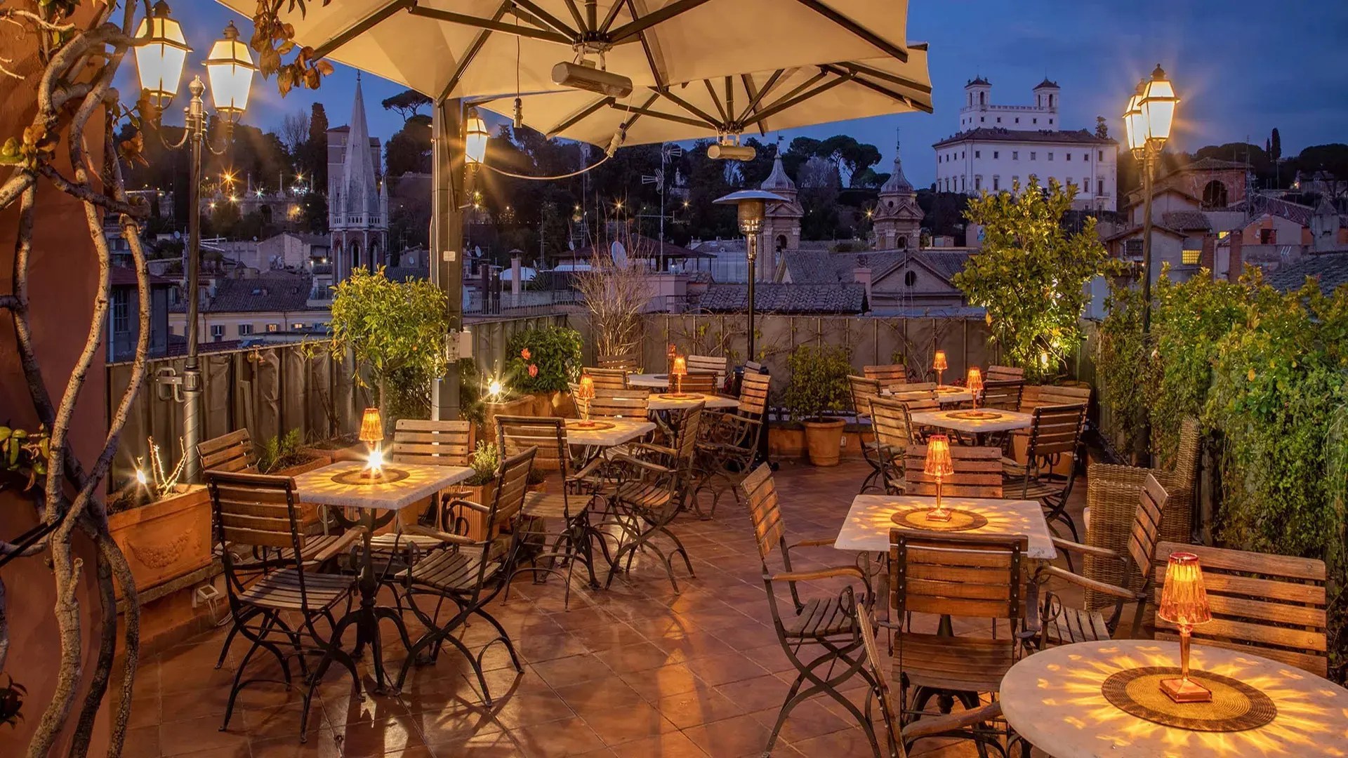 The hotel's rooftop terrace at dusk, with lit candles on set tables, umbrellas and potted plants above the rooftops of Rome.